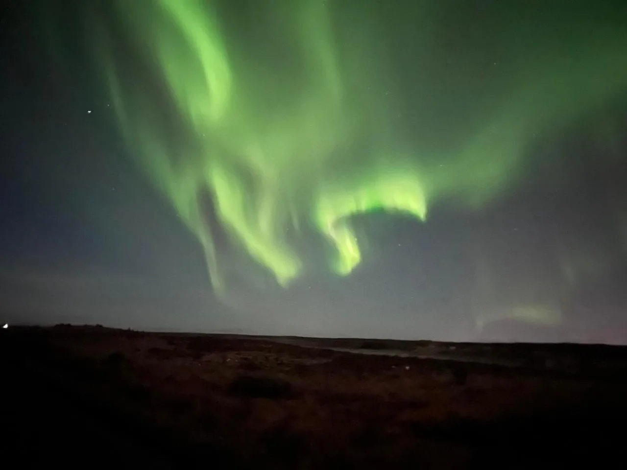 A bright green aurora curtain directly overhead on open ground away from Reykjavik