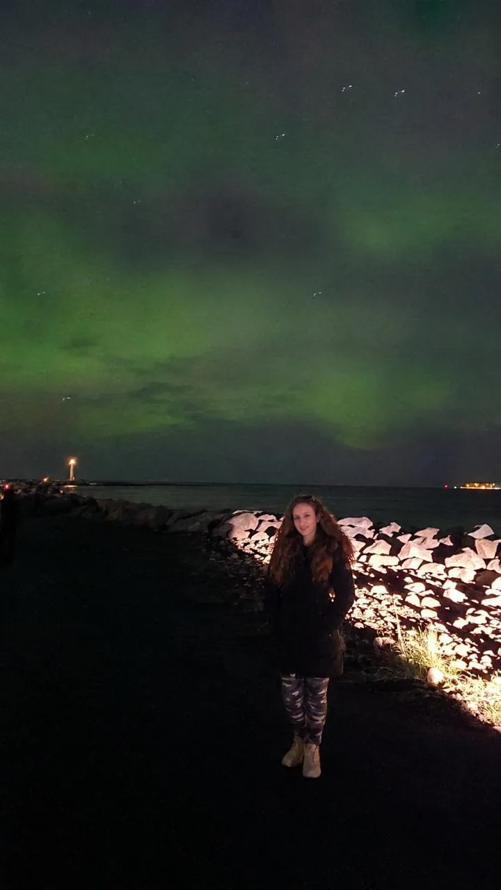 A traveller in a warm coat standing under the northern lights by the lighthouse at Grotta, Reykjavik